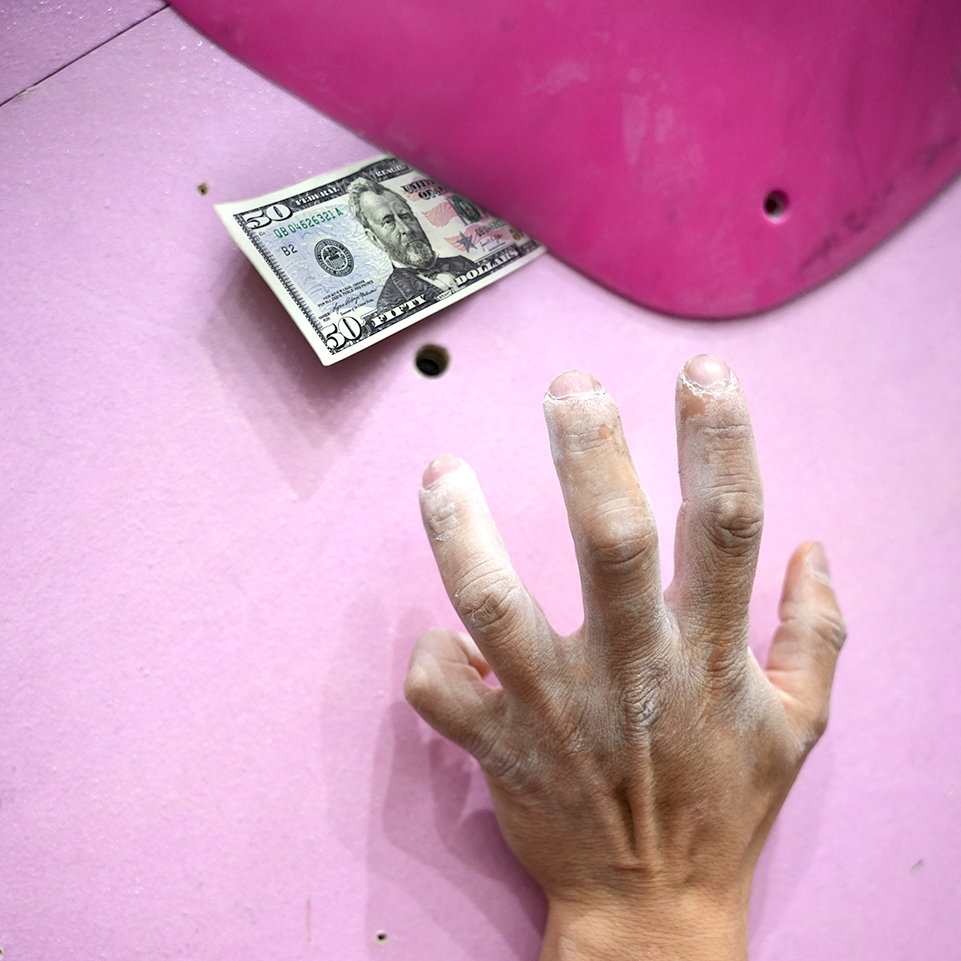 Chalk-covered climber hand reaching toward a hidden $50 bill tucked beneath a pink climbing hold on Sessions’ bounty route