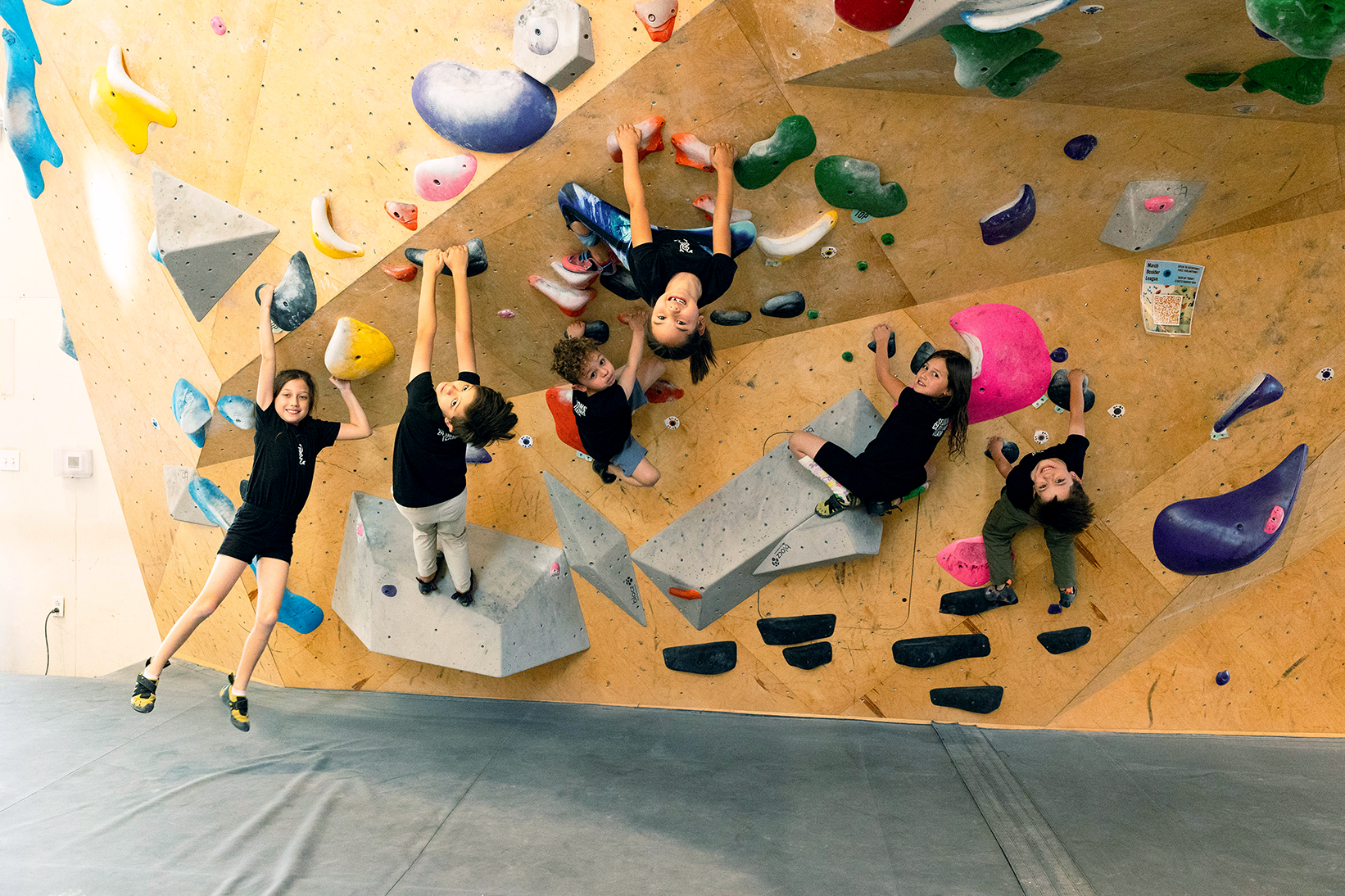 Three young climbers practice bouldering on an indoor climbing wall during a Sessions Tykes climbing class.