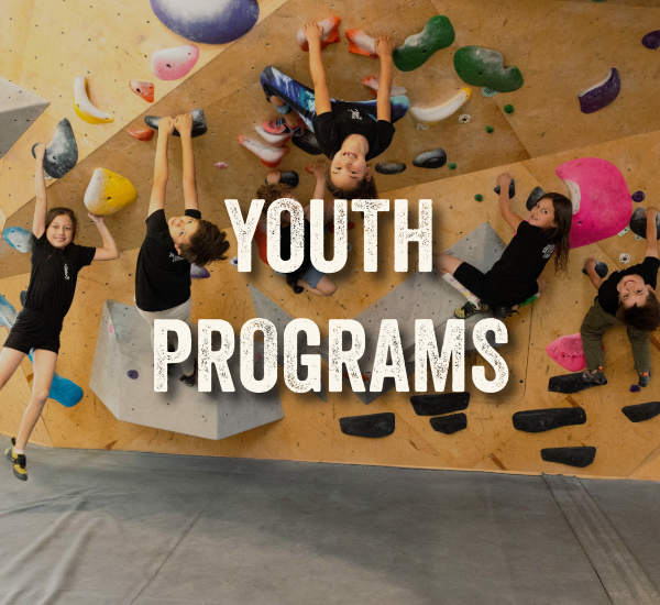 A group of young climbers practice bouldering on an indoor climbing wall during a Sessions Tykes climbing class.